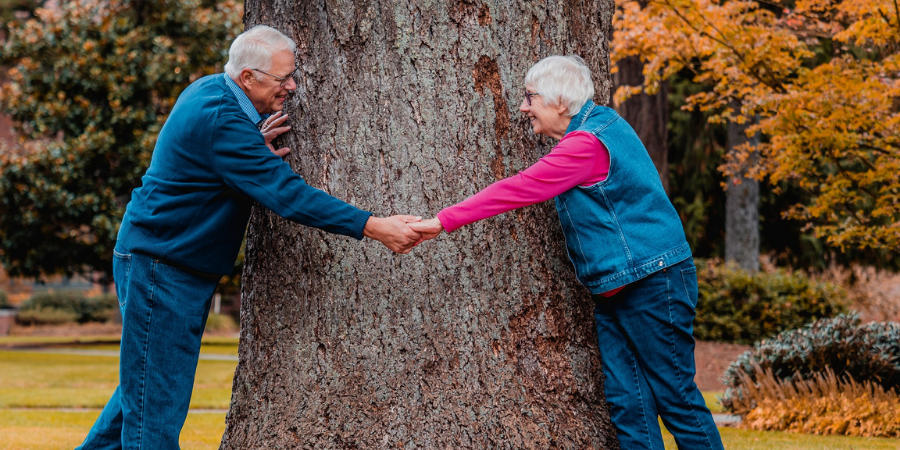 Starszy mężczyzna oraz starsza kobieta stoją bokiem i trzymają się za ręce na tle pnia drzewa.