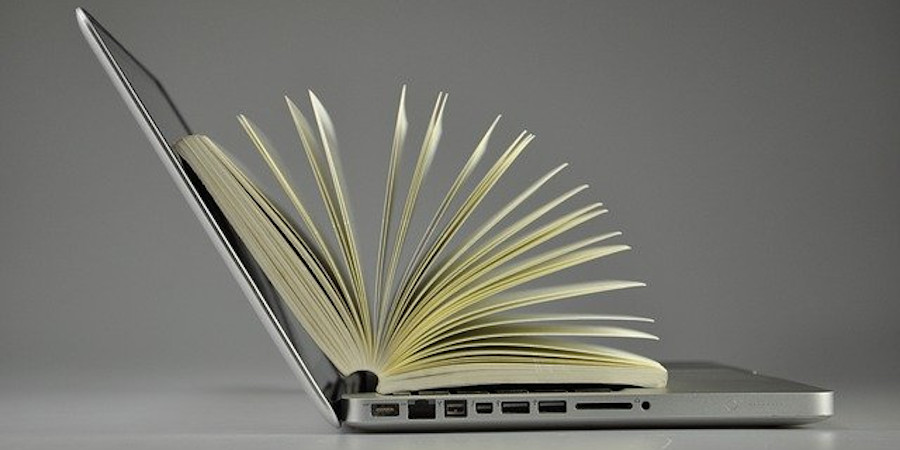 Otwarty laptop, ustawiony do odbiorcy lewym bokiem. Na nim leży otwarta książka, której karty tworzą wachlarz