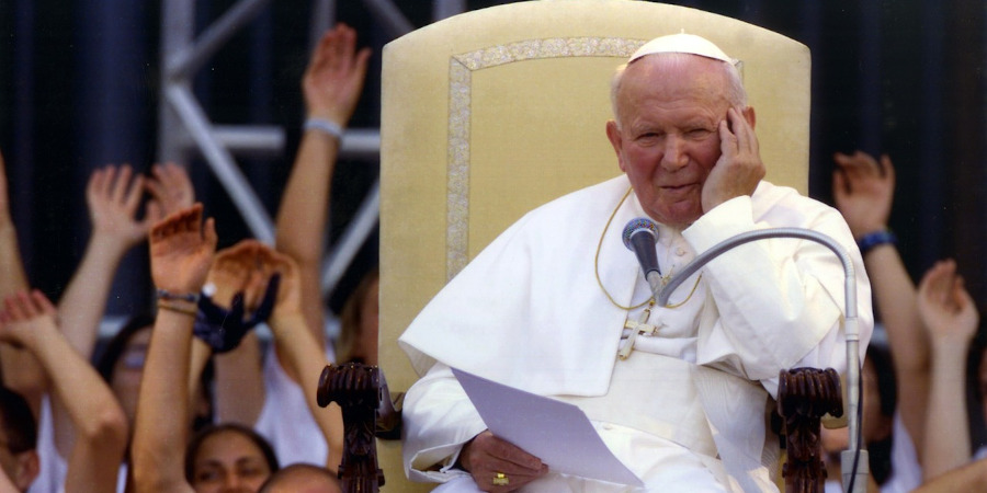 W centralnej części postać Jana Pawła II siedzącego na fotelu, w tle tłum osób wiwatujący rękoma