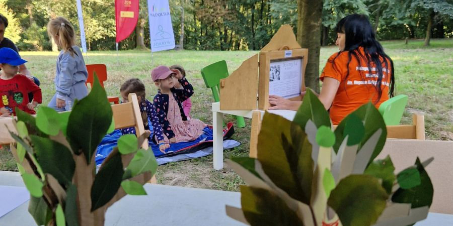 Na pierwszym planie małe kartonowe drzewka stojące na stoliku. Na nich przyklejone zielone liście. W tle kobieta w pomarańczowej koszulce siedząca na krzesełku czytająca bajkę dzieciom siedzącym na kocu. 