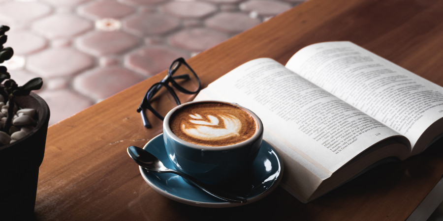 Na drewnianym stoliku otwarta książka, filiżanka z kawą i okulary.