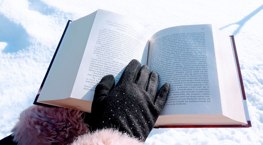 Na tle śniegu otwarta książka trzymana w dłoniach w czarnych skórzanych rękawiczkach.