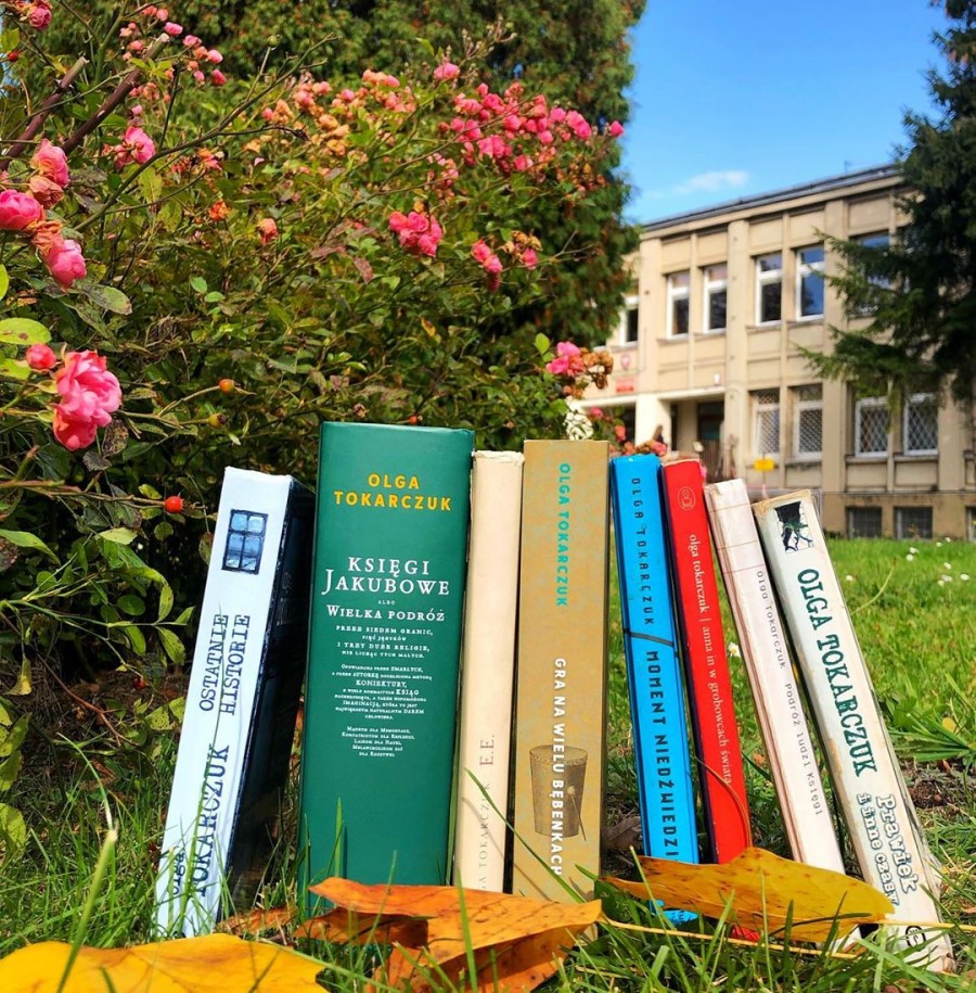 Osiem książek autorstwa Olgi Tokarczuk ułożonych na trawniku przed budynkiem Biblioteki. Od lewej różowe kwiaty piwonii.