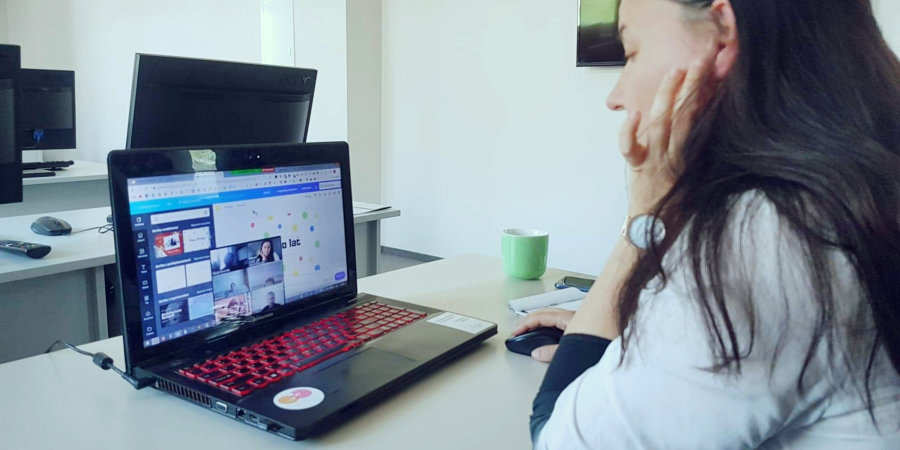 Kobieta siedzi przed komputerem w pustej sali. Na ekranie widać inne osoby, uczestniczące w spotkaniu online.