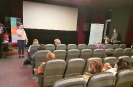 Pracownik Biblioteki Klaudia Kierc stoi z mikrofonem w ręku i przemawia do zebranych w sali kinowej