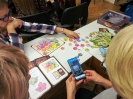 Trzy nauczycielki siedzą przy stoliku nad rozłożoną grą planszową. Jedna z nich zczytuje kod QR z żetonu do gry za pomocą aplikacji w smartfonie.