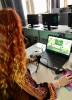 Rudowłosa kobieta siedząca tyłem przy biurku w sali komputerowej. Przed nią laptop, mikrofon oraz notatki. Kobieta trzyma w ręce mysz komputerową