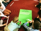 Uczniowie siedzą na dywanie wokół zielonego arkusza z Zasadami Antyhejtowymi do uzupełnienia. Dziewczynka flamastrem dopisuje na karteczce kolejną zasadę.