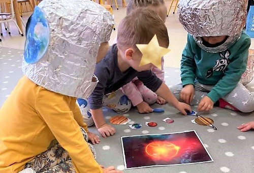 Grupka dzieci w hełmach kosmonautów układa na dywanie ilustracje planet Układu Słonecznego.