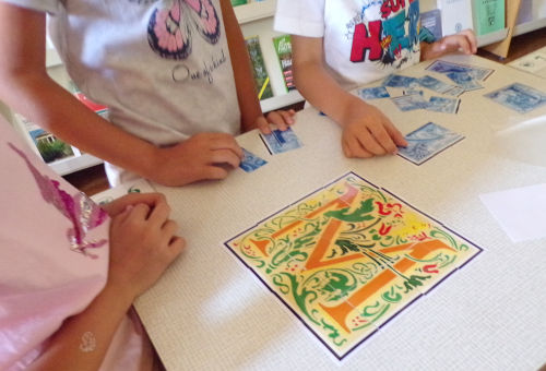 Na stoliku leżący inicjał ułożony z puzzli. Wokół dzieci, które dokładają kolejne elementy.