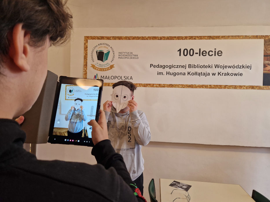 Młody mężczyzna robiący zdjęcie tabletem osobie zasłaniającej twarz maską. Na ścianie za chłopakiem baner z napisem "100-lecie Pedagogicznej Biblioteki Wojewódzkiej im. Hugona Kołłątaja w Krakowie"