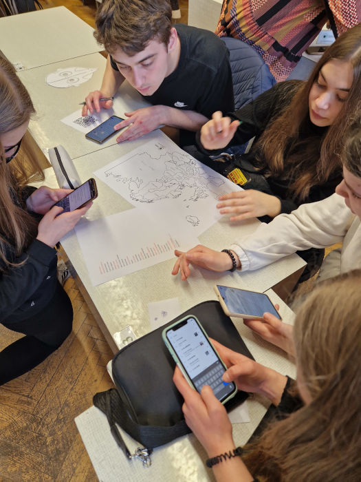 Grupa młodzieży z telefonami w rękach, pochylona nad mapą Europy
