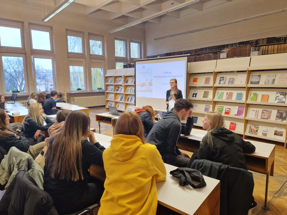 Młoda kobieta tłumaczy zadanie osobom zebranym w sali. W tle slajd z napisem "I Edycja Dnia Autorów Europejskich"