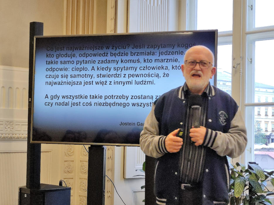 Profesor Grzegorz Leszczyński zwrócony przodem do zgromadzonych osób wygłaszający referat. Za nim wyświetlona na telewizorze prezentacja. 