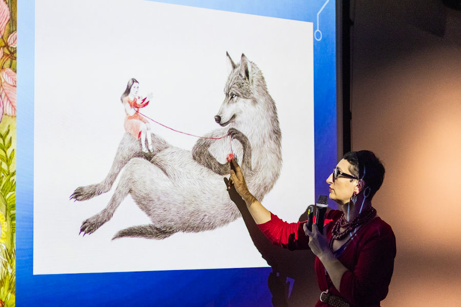 Kobieta w czerwonym ubraniu, z mikrofonem w ręce wskazuje palcami na ekran przedstawiający dziewczynkę i wilka.