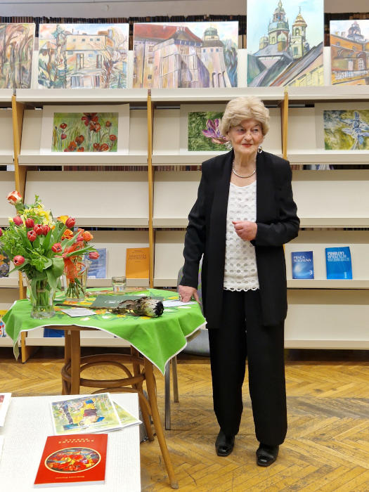 Pani Małgorzata stojąca przy stoliku, w tle obrazy i czasopisma.