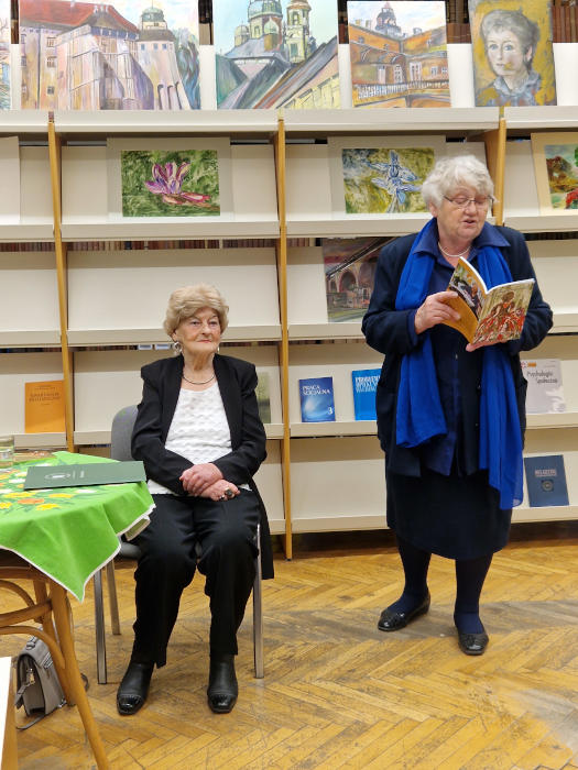 Kobieta w okularach i niebieskim szalu czytająca wiersz Pani Pfisterer, autorka siedzi po lewej stronie.