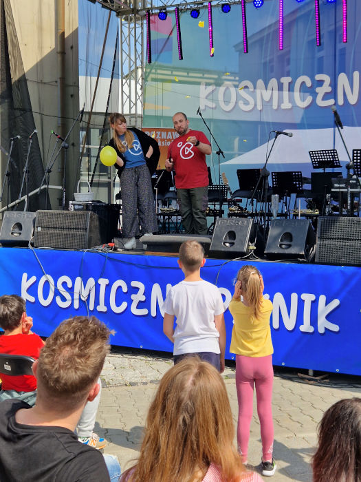 Kobieta i mężczyzna stoją na scenie i pokazują eksperyment z nadmuchanym balonem. Przed sceną stoją dzieci.