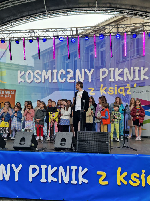 Dzieci z zespołu "Krakowski Chór Dziecięcy" stoi na scenie, trzymając w dłoniach mikrofony.
