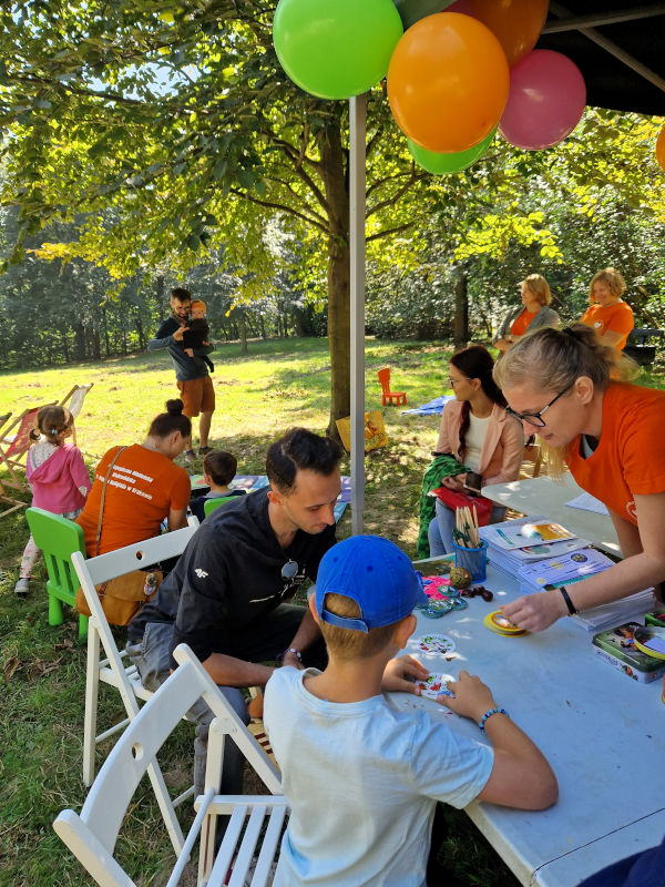 Na pierwszym planie mężczyzna z dzieckiem siedzący przy stoliku grają w grę planszową. Kobieta w pomarańczowej koszulce tłumaczy im zasady. W tle dzieci programujące robota.