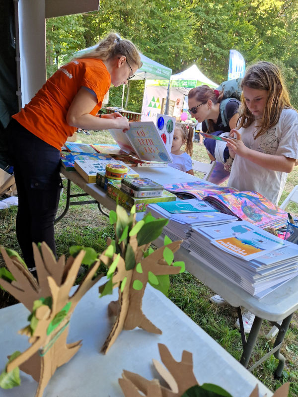 Na pierwszym planie małe kartonowe drzewka z wyciętymi zielonymi liśćmi. W tle kobieta w pomarańczowej koszulce pokazuje książkę osobom przy stoliku.