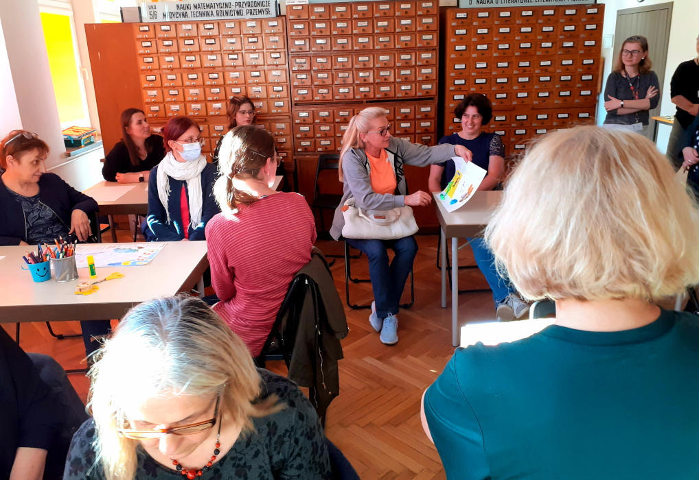 Jedna z uczestniczek pokazuje zebranym przy stolikach kobietom kartkę z ilustracją