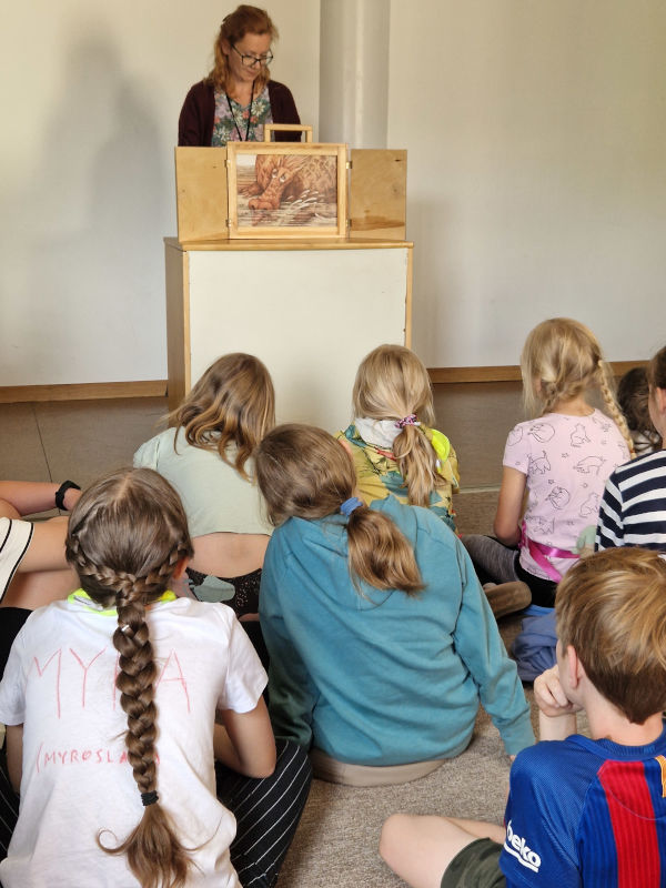 Nauczycielka opowiada dzieciom bajkę korzystając z teatrzyku kamishibai. Dzieci zwrócone w stronę teatrzyku, słuchają opowiadania. 