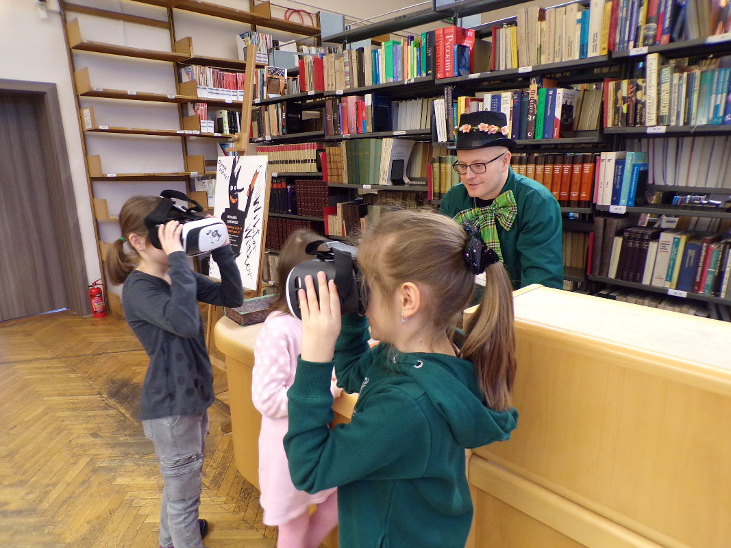 Przy regale z książkami dzieci mają założone na twarzach gogle do wirtualnej rzeczywistości.