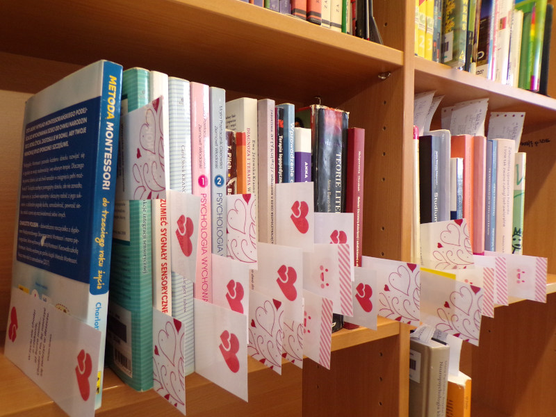 Kolorowe książki ułożone pionowo na regale, a w nich białe zakładki do książek z czerwonymi sercami.
