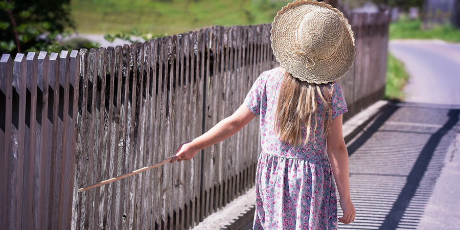 Dziewczynka w letniej sukience i w słomkowym kapeluszu dotyka patykiem drewnianego ogrodzenia.
