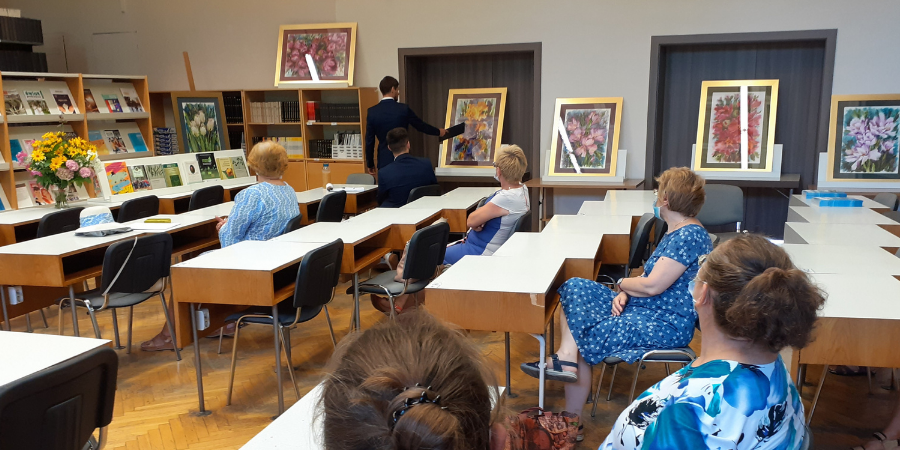 Grupa osób siedzących przy stolikach patrzy na wystawę obrazów przedstawiających kwiaty.