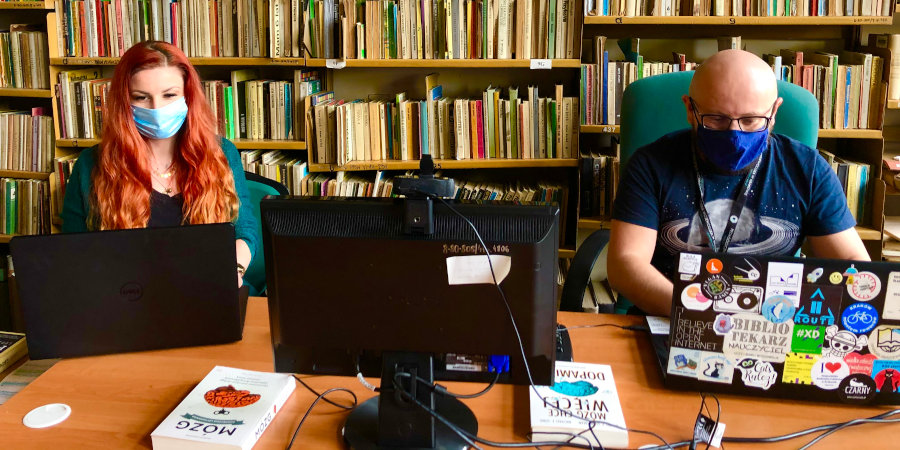 Na tle bibliotecznych regałów siedzą przed ekranami komputerów - po lewej Marta Jesionek, po prawej Karol Baranowski - nauczyciele bibliotekarze z naszej biblioteki.