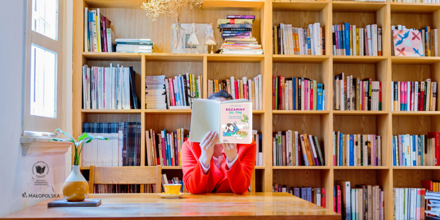 Kobieta trzymająca w rękach książkę "Egzaminy bez spiny", w tle regały z książkami. W lewym dolnym rogu logo Biblioteki.