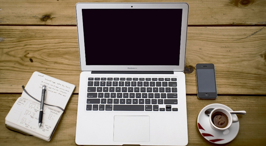 Tło z desek, na środku otwarty laptop, po lewej stronie zapisany notatnik z piórem, po prawej smartfon i filiżanka z czarną kawą. 