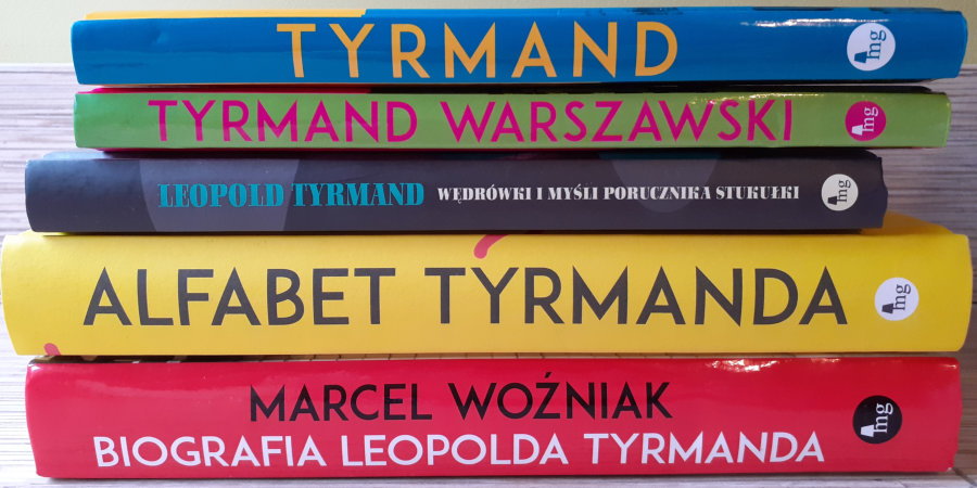 Na jasnym stole, na tle jasnej ściany leży 5 kolorowych grzbietów książek Leopolda Tyrmanda i o Leopoldzie Tyrmandzie.