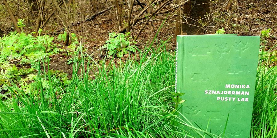 W prawym dolnym rogu zielona okładka książki "Pusty las" Moniki Sznajderman. W tle zielona wiosenna trawa i brązowe pnie oraz gałęzie drzew