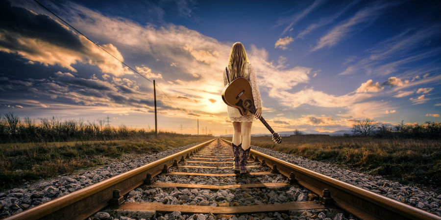 Blondynka z gitarą zmierza środkiem torów kolejowych w stronę zachodzącego słońca.