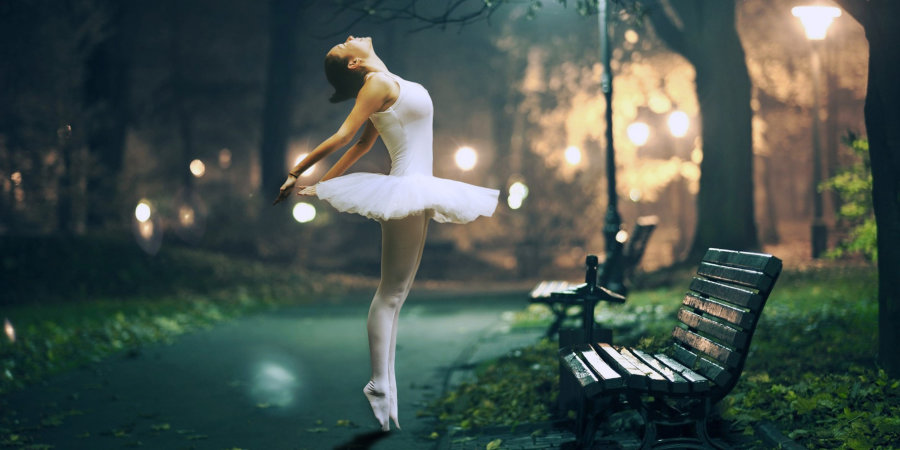 Baletnica w białym klasycznym stroju, obrócona bokiem podskakuje na chodniku nocą. Obok tancerki drewniana ławka i zapalona latarnia. Zielone liście wokół chodnika.