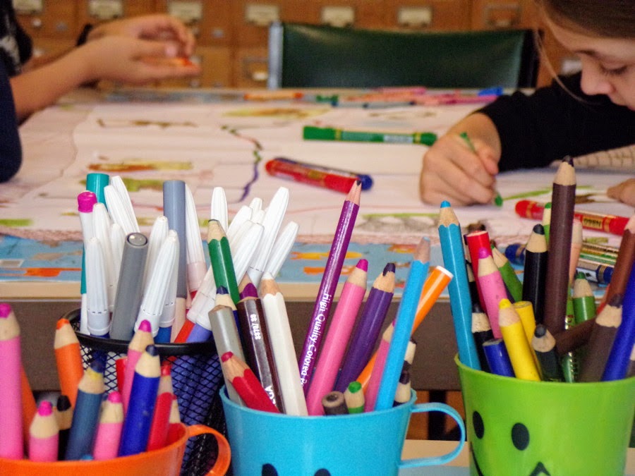 Kolorowe kredki w plastikowych kubeczkach, za nimi ręka rysującego dziecka.