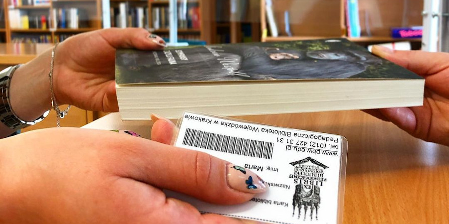 Dwie damskie dłonie nad ladą w bibliotecznej wypożyczalni podają sobie równocześnie z jednej strony książkę, z drugiej kartę biblioteczną czytelniczki