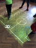Na podłodze z projektora Funtronic wyświetlone jest pomniejszone, zielone boisko do piłki nożnej. Chłopiec w zielonej bluzie kopie wirtualną biało-czarną piłkę.