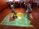 Dwoje uczniów gra w rozgrywki piłkarskie na Magicznym Dywanie, w tle dopingują koledzy i koleżanki