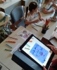 Prowadząca warsztaty trzyma w ręku tablet z aplikacją Quiver, w tle kolorowanki dzieci i kredki