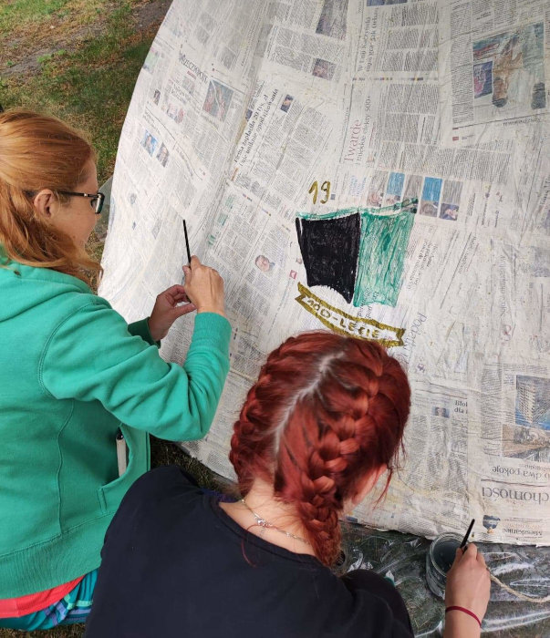 Małgorzata Stachurska i Marta Jesionek malują farbami jubileuszowe logo Pedagogicznej Biblioteki Wojewódzkiej w Krakowie na szałasie zrobionym z gazet