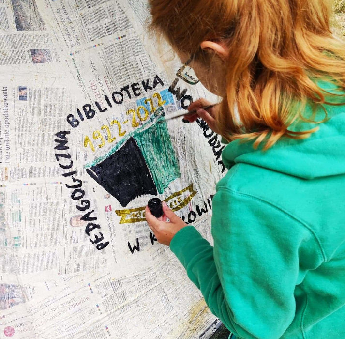 Małgorzata Stachurska uzupełnia detale w logo Biblioteki malowanym na szałasie z gazet