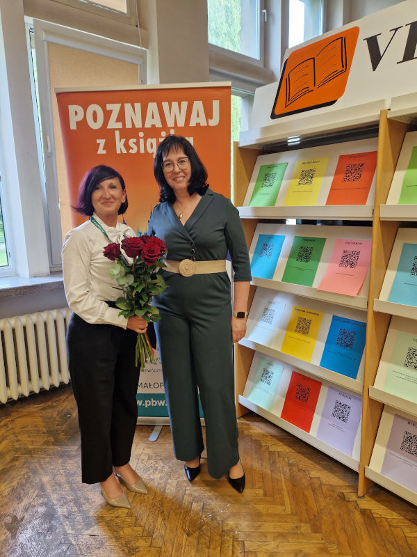 Doktor Dagmara Kawoń-Noga oraz Pani Dyrektor Anna Piotrowska zwrócone przodem do obiektywu, trzymające czerwone róże na tle półek z czasopismami. 