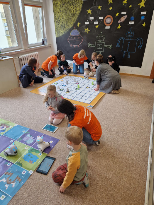 Przy matach z robotami Photonami ,na dywanie siedzą dzieci i kobiety w pomarańczowych koszulkach. Dzieci mają przed sobą tablety i programują roboty.