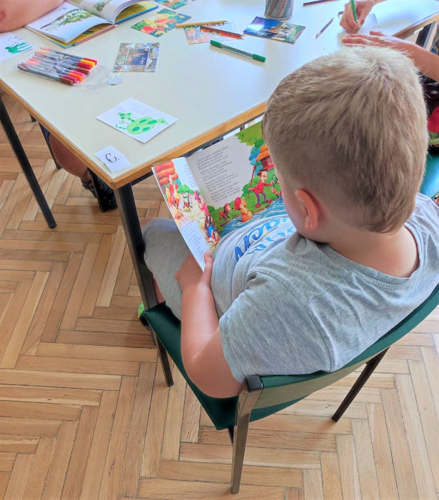 Chłopiec czytający książkę, przed nim stół na którym rozłożone są pocztówki, pisaki i kredki