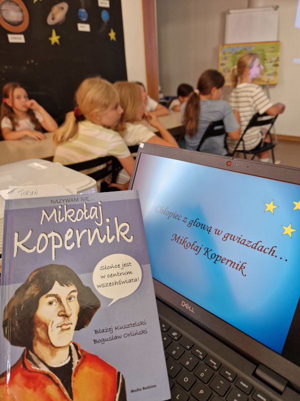 Na pierwszym planie książka o Mikołaju Koperniku leżąca przy komputerze. Z tyłu dzieci oglądające prezentację.