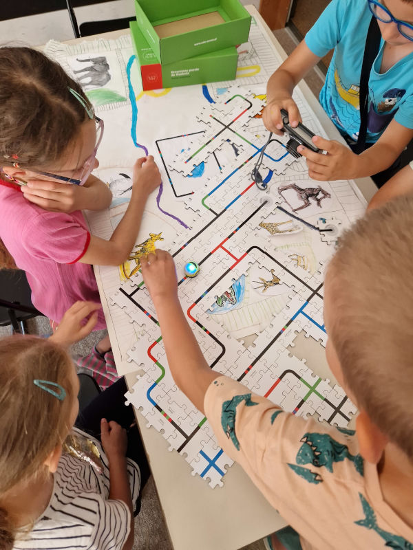 Grupa dzieci układających kolorowe, drewniane puzzle. Na nich jeżdżący mały robot. 